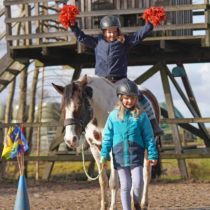 Zwei Kinder jubeln mit Pompons auf dem Kletterturm, darunter wartet ein Pony